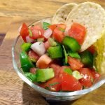 A bowl of pico de gallo with tomato, green peppers, jalapeño, and cilantro topped with two tortilla chips sitting on a wooden cutting board.