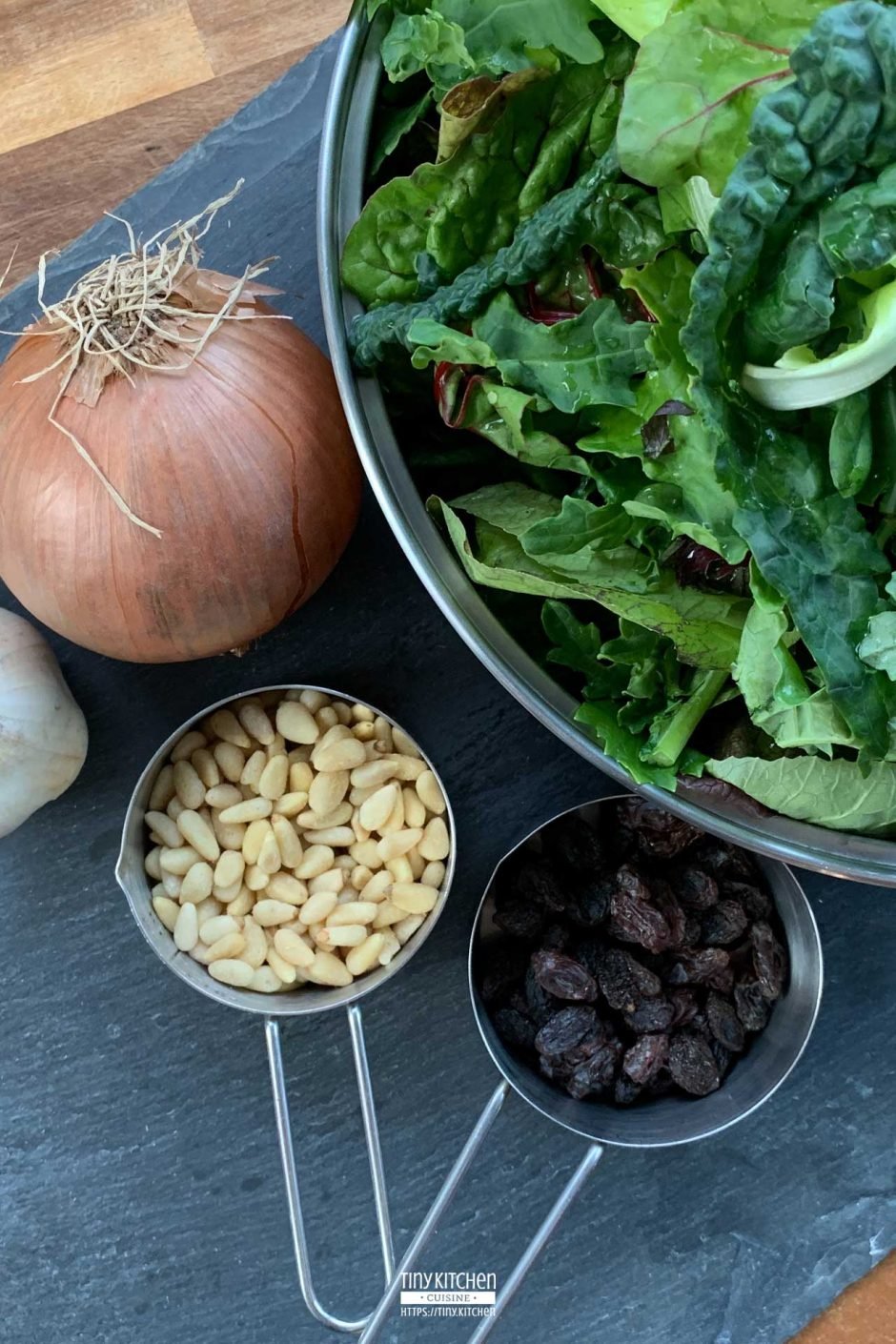 A bowl of fresh mixed braising greens next to an onion, a head of garlic, and two measuring cups filled with pine nuts and raisins.