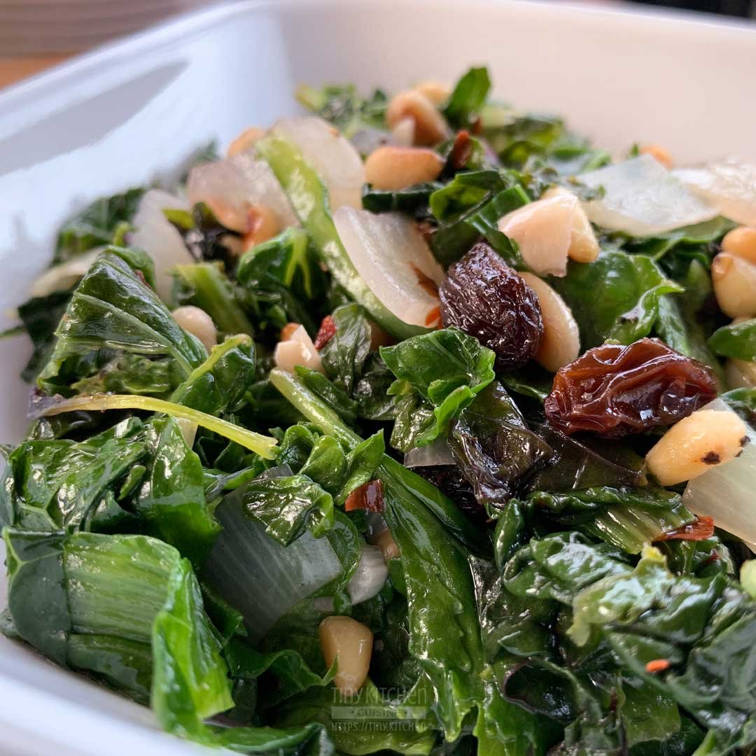 Closeup of a bowl of mixed braised greens topped with toasted pine nuts and raisins.