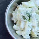 A metal bowl of creamy cucumber salad with fresh dill sitting on a slate board.